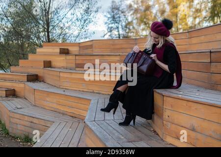 Une belle femme est assise sur un banc de théâtre d'été en bois dans un manteau bordeaux et le biret, regarde la caméra, à l'automne contre le ciel bleu. Banque D'Images