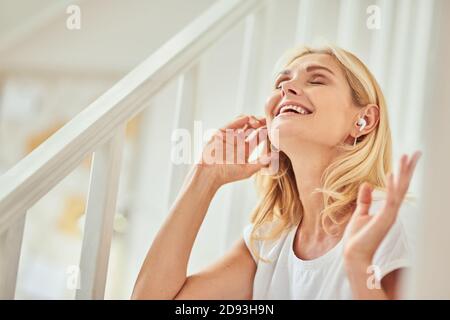 Bonne femme de race blanche d'âge moyen appréciant la musique avec les écouteurs sans fil tout en étant assise à l'intérieur avec les mains levées et les yeux fermés. Les gens passent du temps à la maison Banque D'Images