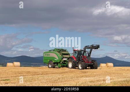 Un tracteur Valtra rouge et une presse à balles McHale Fusion Vario dans un champ avec balles de paille. Les collines de Menaway et Bennachie sont visibles au loin. Banque D'Images