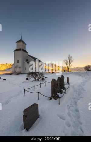 La petite église blanche en bois de Gimsøy sur la plage Sur les îles Lofoten en Norvège en hiver avec belle vieux cimetière et montagnes le soir l Banque D'Images