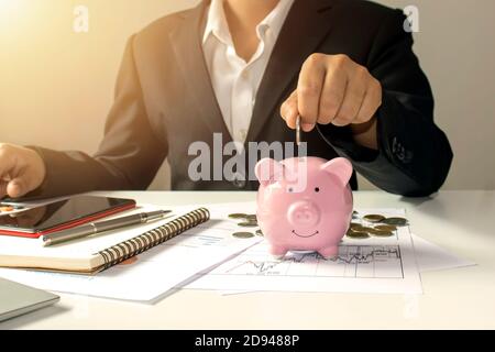 Une femme d'affaires qui met des pièces de monnaie dans une banque de pigey, y compris un carnet sur un bureau en bois pour le travail, des idées pour économiser de l'argent et la gestion de comptes. Banque D'Images