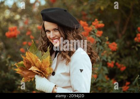 Bonjour septembre. Femme souriante et branchée de 40 ans en trench beige et béret noir avec feuilles jaunes d'automne dehors sur la rue de la ville en automne Banque D'Images