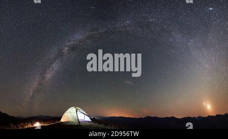 Vue panoramique du camping touristique la nuit dans les montagnes. Tente illuminée et feu de camp sous un beau ciel nocturne plein d'étoiles et de voie lactée Banque D'Images