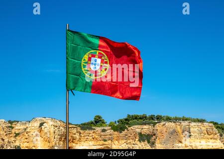 Accent sélectif sur l'agissement du drapeau du portugal devant la formation typique de roche jaune de l'algarve et du ciel bleu. Drapeau national du portugal avec espace. Banque D'Images