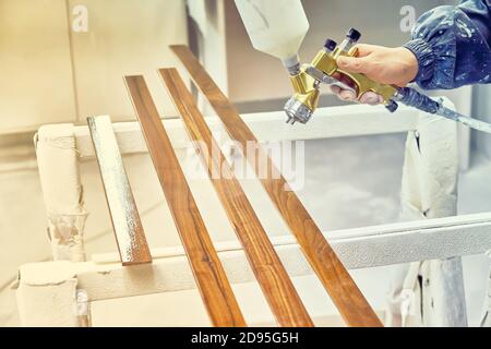 Homme peignant des détails de mobilier dans une cabine de pulvérisation. Travailleur utilisant un pistolet pulvérisateur. Fabrication de meubles. Procédé de fabrication de meubles en bois. Image en tons Banque D'Images