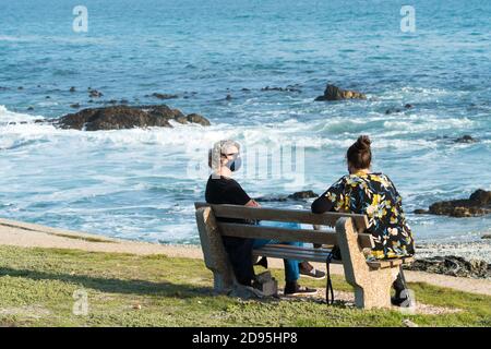 deux femmes discutent sur un banc surplombant l'océan ou de la mer pendant une pandémie portant des masques faciaux et de maintien un virus corona de concept de distance de sécurité Banque D'Images