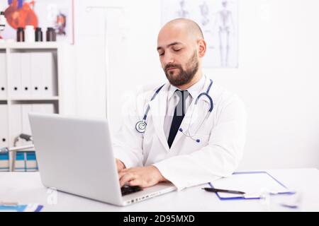 Médecin portant un stéthoscope et un manteau blanc dans le bureau de l'hôpital utilisant un ordinateur portable pour écrire le traitement du patient. Médecin utilisant le bloc-notes dans le milieu de travail de la clinique , confiant, expertise, médecine. Banque D'Images