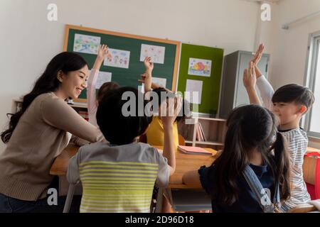 Jeune femme travaillant à l'école avec le bras levé, les élèves mettant leurs mains vers le haut pour répondre à la question, enthousiasme, désireux, plaisir. Professeur d'école asiatique avec des élèves qui élèvent la main Banque D'Images