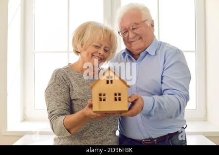 Couple senior souriant tenant une maison miniature debout près de la fenêtre dans leur nouvelle maison Banque D'Images