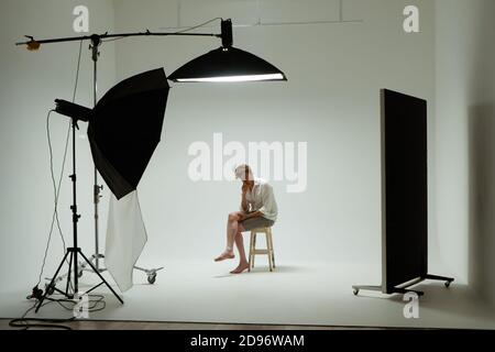 Un jeune homme pieds nus est assis sur une chaise en posture de penseur posé en studio photo. Modèle masculin portant une chemise blanche sur fond blanc parmi les équipements de studio Banque D'Images