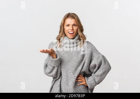 Image de la femme blonde frustrée et embêtue dans le chandail gris besoin de réponses, levant la main et frowning, debout sur fond blanc Banque D'Images