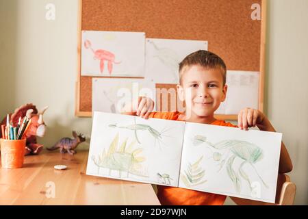 Adorable enfant garçon dessin dinosaures sur la photo avec des crayons de couleur assis près d'un bureau dans sa chambre Banque D'Images