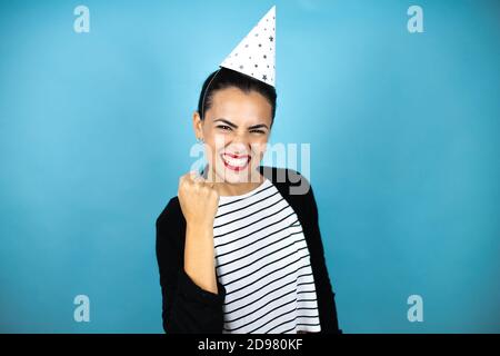 Jeune belle femme portant un chapeau d'anniversaire sur bleu insolé contexte en colère et fou soulevant poing frustré et furieux pendant crier avec ange Banque D'Images