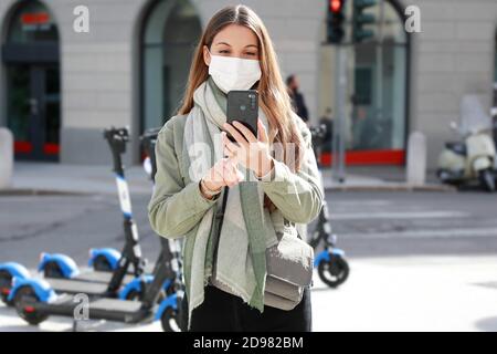 Une jeune femme avec masque de protection télécharge l'application à utiliser le scooter électrique dans la rue de la ville Banque D'Images