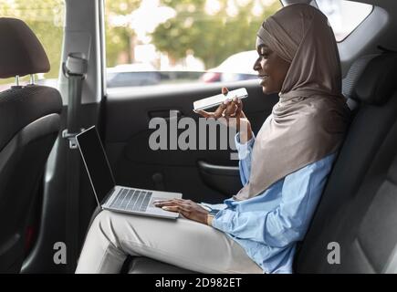 Femme musulmane noire envoyant un message vocal et utilisant l'ordinateur portable dans Voiture Banque D'Images