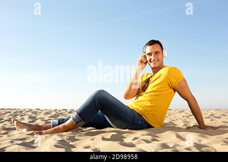 Portrait d'un homme souriant pieds nus assis dans le sable au plage écouter de la musique avec un casque Banque D'Images