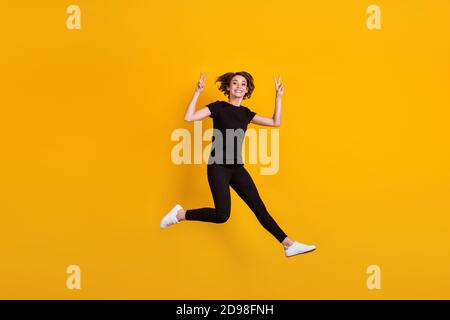 Vue sur toute la longueur du corps de la jolie fille gaie funky saut de plaisir montrant le v-signe isolé couleur jaune vif arrière-plan Banque D'Images