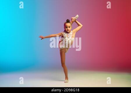 Énergie. Petite fille caucasienne, formation de gymnaste rhytmique, exécutant isolé sur fond dégradé bleu-rouge studio au néon. Enfant gracieux et souple, fort. Concept de sport, de mouvement, d'action. Banque D'Images