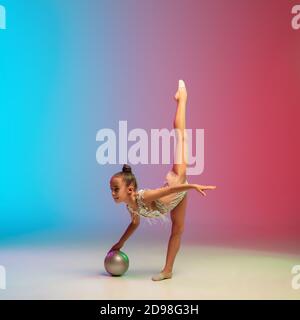 Énergie. Petite fille caucasienne, formation de gymnaste rhytmique, exécutant isolé sur fond dégradé bleu-rouge studio au néon. Enfant gracieux et souple, fort. Concept de sport, de mouvement, d'action. Banque D'Images