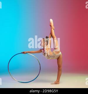 Mouvement. Petite fille caucasienne, formation de gymnaste rhytmique, exécutant isolé sur fond dégradé bleu-rouge studio au néon. Enfant gracieux et souple, fort. Concept de sport, de mouvement, d'action. Banque D'Images