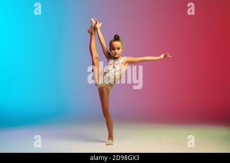 Dynamique. Petite fille caucasienne, formation de gymnaste rhytmique, exécutant isolé sur fond dégradé bleu-rouge studio au néon. Enfant gracieux et souple, fort. Concept de sport, de mouvement, d'action. Banque D'Images