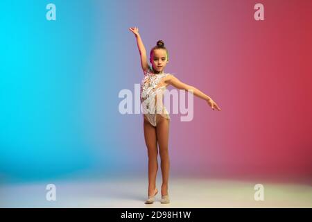 Dynamique. Petite fille caucasienne, formation de gymnaste rhytmique, exécutant isolé sur fond dégradé bleu-rouge studio au néon. Enfant gracieux et souple, fort. Concept de sport, de mouvement, d'action. Banque D'Images