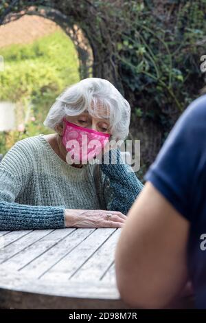 Femme mature portant un masque rendant visite à la mère sénior Lonely dépressive Jardin pendant le verrouillage Banque D'Images