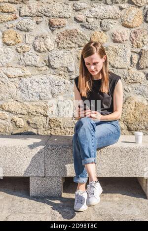 belle jeune femme habillant des vêtements décontractés et assise sur un banc de granit tout en utilisant son smartphone noir un jour ensoleillé, projetant son ombre Banque D'Images