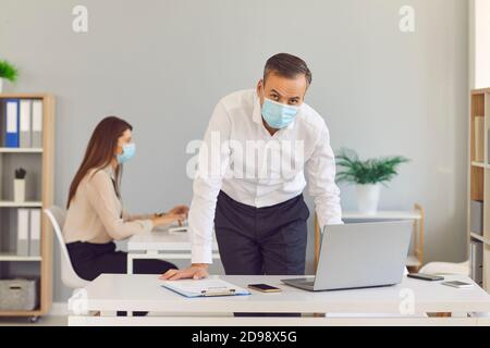 L'homme adulte dans un masque médical de protection se tient dans le bureau contre le fond de son collègue. Banque D'Images