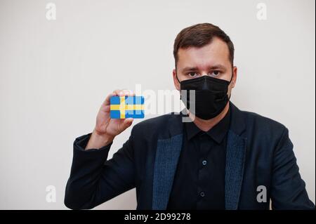 L'homme européen porte un masque classique noir et protège le visage, tenir la carte drapeau de Suède isolée sur fond blanc. Europe coronavirus Covid pays concept. Banque D'Images