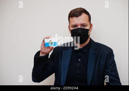 L'homme européen porte noir formel et protège masque, tenir la carte drapeau de Saint-Marin isolée sur fond blanc. Europe coronavirus Covid pays concept Banque D'Images