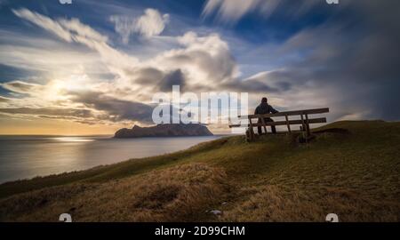 Homme sur banc au village de Gasadalur et à l'île de Mykines au coucher du soleil, îles Féroé Banque D'Images