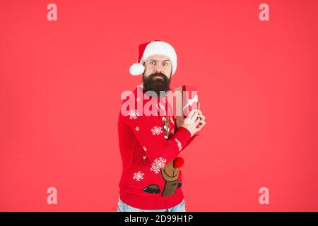 un gars à barbe cupide portant un pull tricoté amusant et un chapeau du père noël et une boîte à présent a une idée pour célébrer la fête du nouvel an, le jour de l'an de noël. Banque D'Images