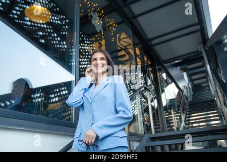 Femme gaie portant un costume bleu habillé debout devant les fenêtres de bureau sur fond regardant vers le haut confiant et souriant plein d'espoir, arranger les cheveux à la main, c Banque D'Images