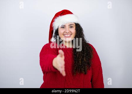 Jeune belle femme portant un chapeau de père Noël sur fond blanc souriant amical offrant poignée de main comme accueil et accueillant Banque D'Images