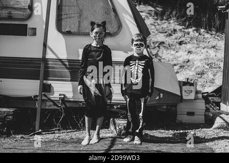 Photo en niveaux de gris d'une jolie sœur et d'un frère portant des costumes d'Halloween et debout contre un campeur Banque D'Images