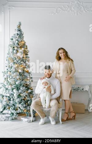 Le concept d'un Noël en famille. Une belle famille en vêtements légers est assise à côté d'un arbre de Noël décoré. Famille heureuse avec un bébé sur un ch Banque D'Images