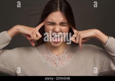 Une jeune femme dans une situation stressante presse ses temples avec ses doigts et hurle. Photo de studio sur fond sombre. Banque D'Images