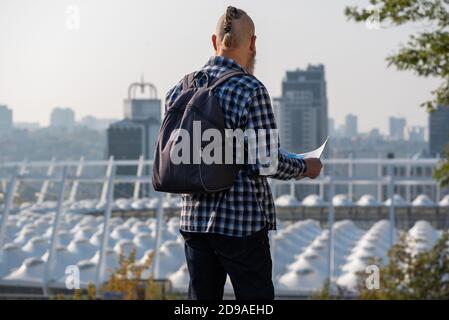 Homme caucasien barbu touriste en chemise à carreaux avec sac à dos sur son dos regardant la carte de planification de l'itinéraire dans le centre-ville. Vue arrière Banque D'Images