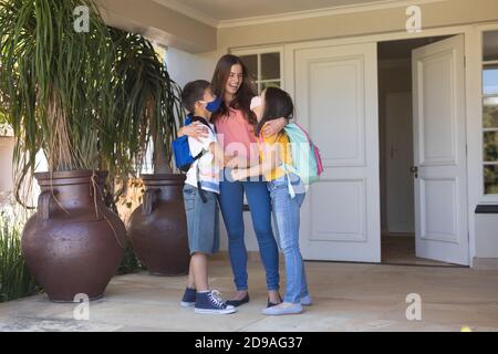 Un garçon et une fille de race blanche portant un masque facial embrassant leur mère à la maison avant de partir pour l'école Banque D'Images