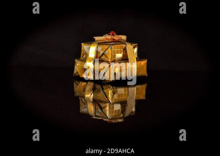 Petits paquets de cuboïdes cadeau enveloppés dans du papier d'emballage doré et ruban or brillant, isolés sur une surface réfléchissante noire, montrant un mirro inversé Banque D'Images