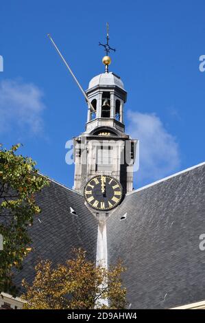 Une tour d'horloge en ardoise avec des chiffres romains à midi. La tour de l'horloge est à la base d'un clocher à un sommet d'angle sur un toit en ardoise carrelée. Banque D'Images