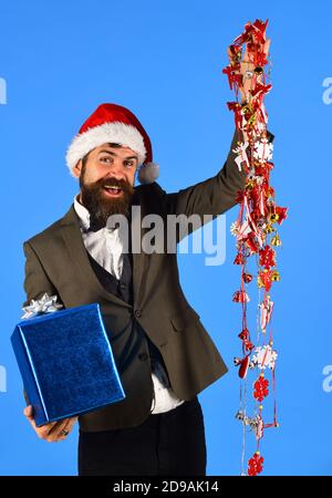 Homme d'affaires avec le visage joyeux tient boîte et décorations. Le Manager avec barbe tient un cadeau bleu. Homme en costume élégant, chapeau de père Noël et guirlande sur fond bleu. Concept d'affaires et de célébration. Banque D'Images
