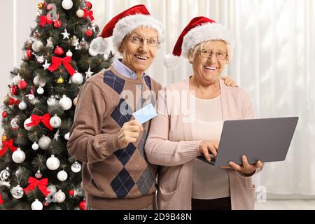 Couple de personnes âgées faisant des achats de noël en ligne avec un ordinateur portable et carte de crédit dans une chambre avec un arbre de noël Banque D'Images