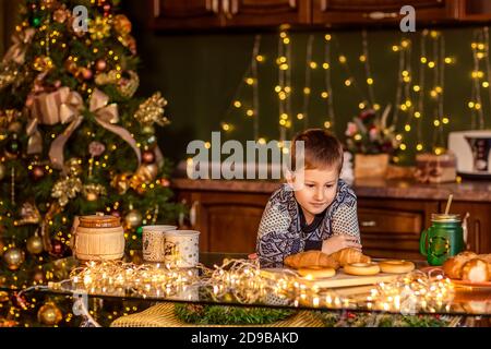 Un garçon est assis à une table dans une cuisine de Noël décorée. Il regarde le croissant et veut le manger. Soirées d'hiver confortables à la maison. Concept du Christ Banque D'Images