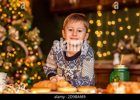 Un garçon est assis à une table dans une cuisine de Noël décorée. Il regarde le croissant et veut le manger. Soirées d'hiver confortables à la maison. Concept du Christ Banque D'Images