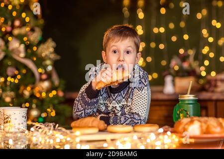 Un garçon est assis à une table dans une cuisine de Noël décorée. Il regarde le croissant et veut le manger. Soirées d'hiver confortables à la maison. Concept du Christ Banque D'Images