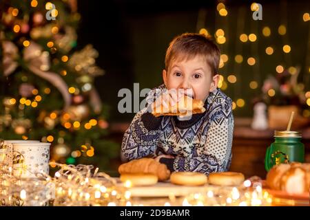 Un garçon est assis à une table dans une cuisine de Noël décorée. Il regarde le croissant et veut le manger. Soirées d'hiver confortables à la maison. Concept du Christ Banque D'Images