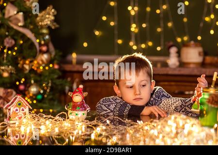 Un garçon est assis à une table dans une cuisine de Noël décorée. Tenir un jouet pour décorer un arbre de Noël. Soirées d'hiver confortables à la maison. Concept du Christ Banque D'Images