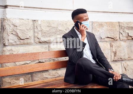 Homme d'affaires portant un masque assis à l'extérieur sur un banc avec téléphone ayant Pause déjeuner pendant une pandémie Banque D'Images